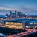 Blue-hour panoramic view of Rogers Place, the Edmonton Convention Centre, the Edmonton EXPO Centre, and Commonwealth Stadium with warm interior lights and a softly blurred downtown skyline.